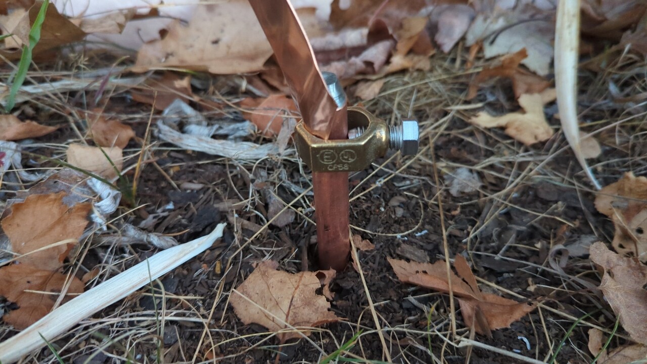 Top of a ground rod peeking above ground level with a screw clamp and a copper ribbon connected to it