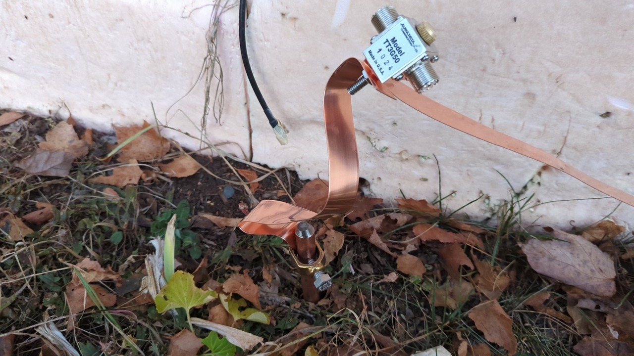 Inline surge protector hanging from its copper ribbons near a ground-level window; one ribbon connects to a ground rod and the other extends off to the right