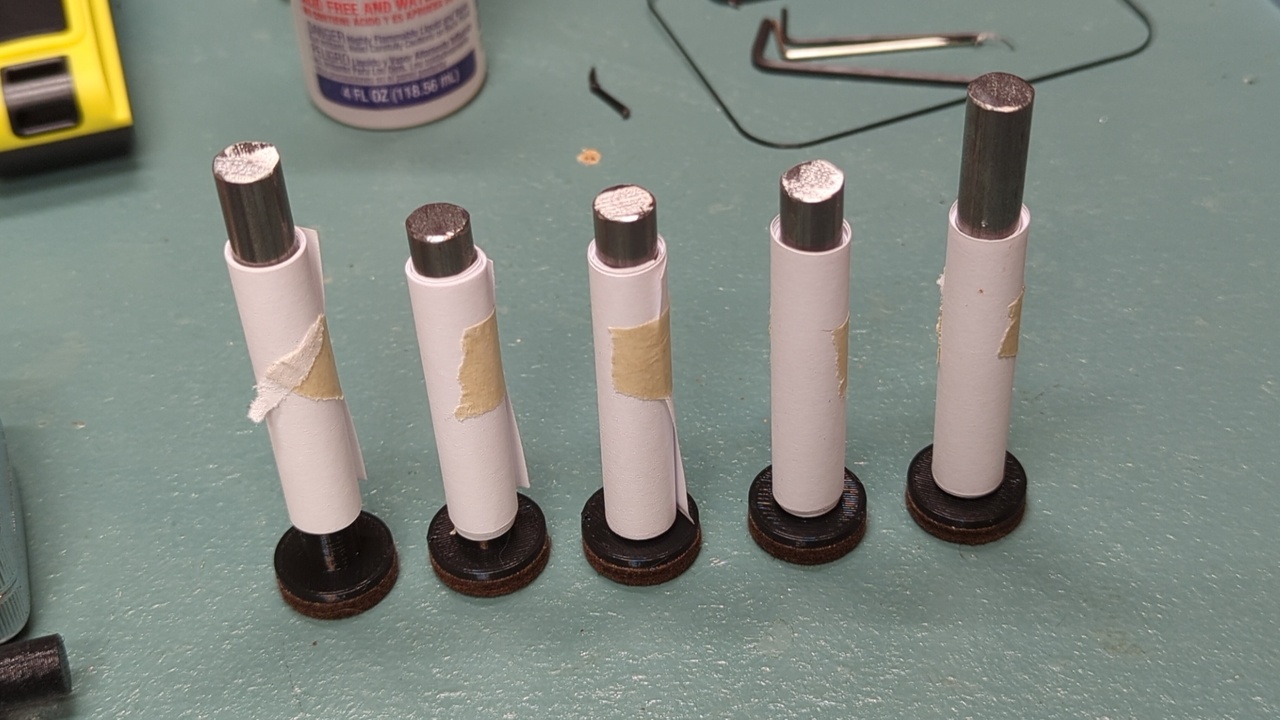 Five plungers and their extensions sitting on a table with paper rolls stabilizing them while their glue dries