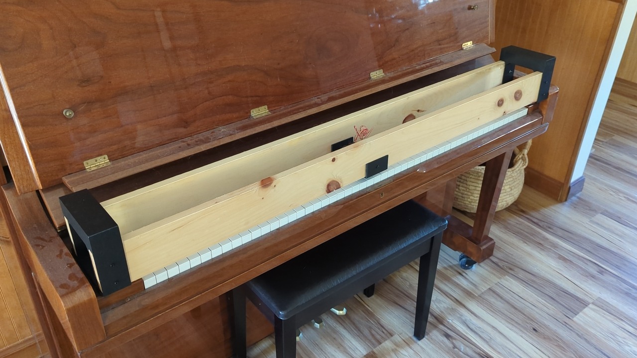 Two wooden boards span the length of an upright piano keyboard. Black plastic support structures connect the boards to the piano on either end.