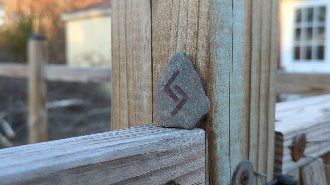 A small, flat stone rests on top of a fence railing and against one of the posts. It has a symbol drawn in dark ink that looks like a less-than and a greater-than sign facing each other, except they are offset so their “jaws” interlock.