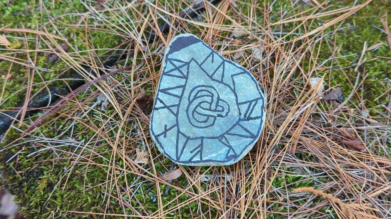 A rock with a flat face rests on a bed of moss and pine needles on top of a stone wall. The rock has several symbols drawn around its edge in dark ink, and a knot is drawn in the center.