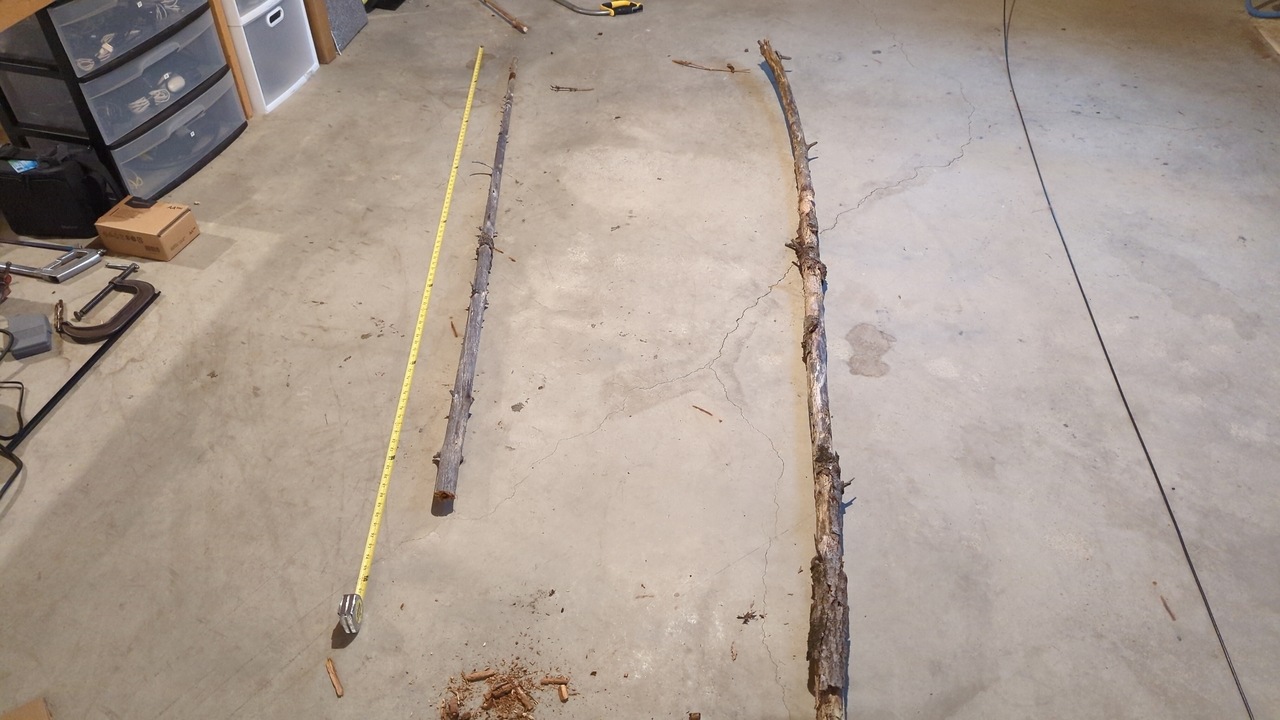 Two sticks lay on a concrete floor with some tools nearby. A tape measure extends parallel to them and indicates that they are about seven feet long.
