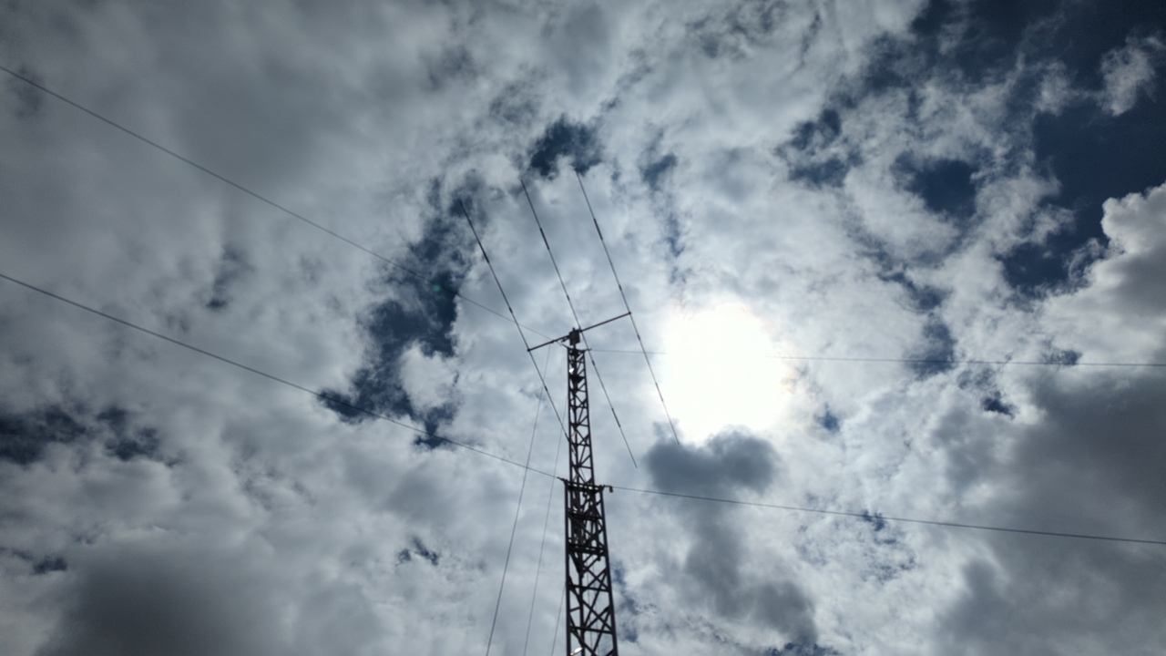 Beam antenna atop an aluminum tower with guy wires visible and the sun and broken clouds above