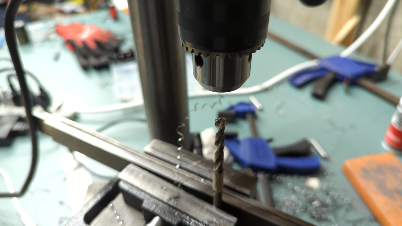 Drill press with empty chuck and a drill bit clamped in a vise. Chips have fallen everywhere, and other tools, gloves, and cables are visible in the background.