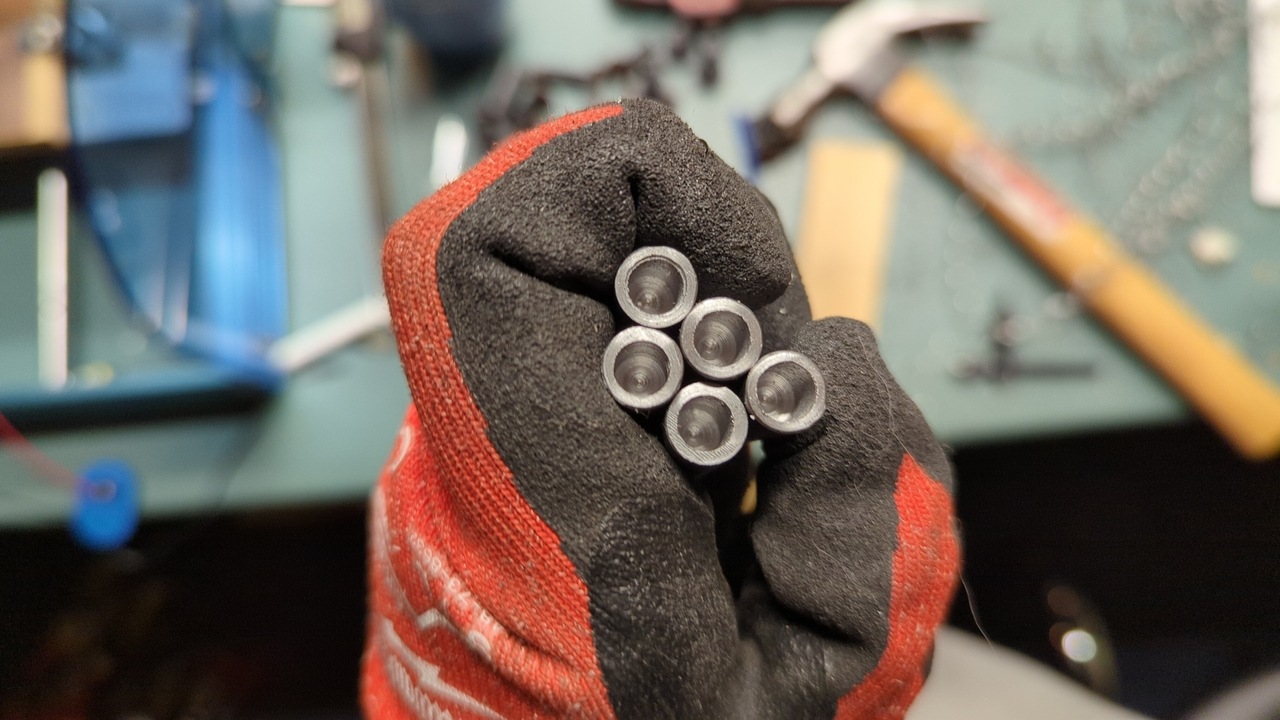 Five short steel rods with centered holes drilled in one end. A gloved hand holds the rods in a bundle viewed from above.