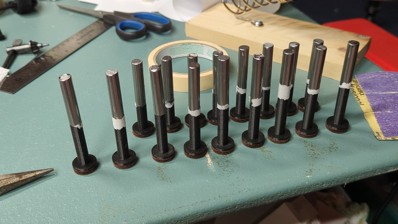 Seventeen plungers of the old design standing together on a table. Most of the plungers have some ripped bits of paper stuck to them at the interface between the steel and plastic parts.
