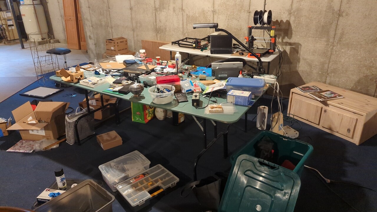 My lab at the beginning of this project. Tables are completely filled with parts, tools, boxes, and junk. The tables are surrounded by similar piles, and the lights are dim.